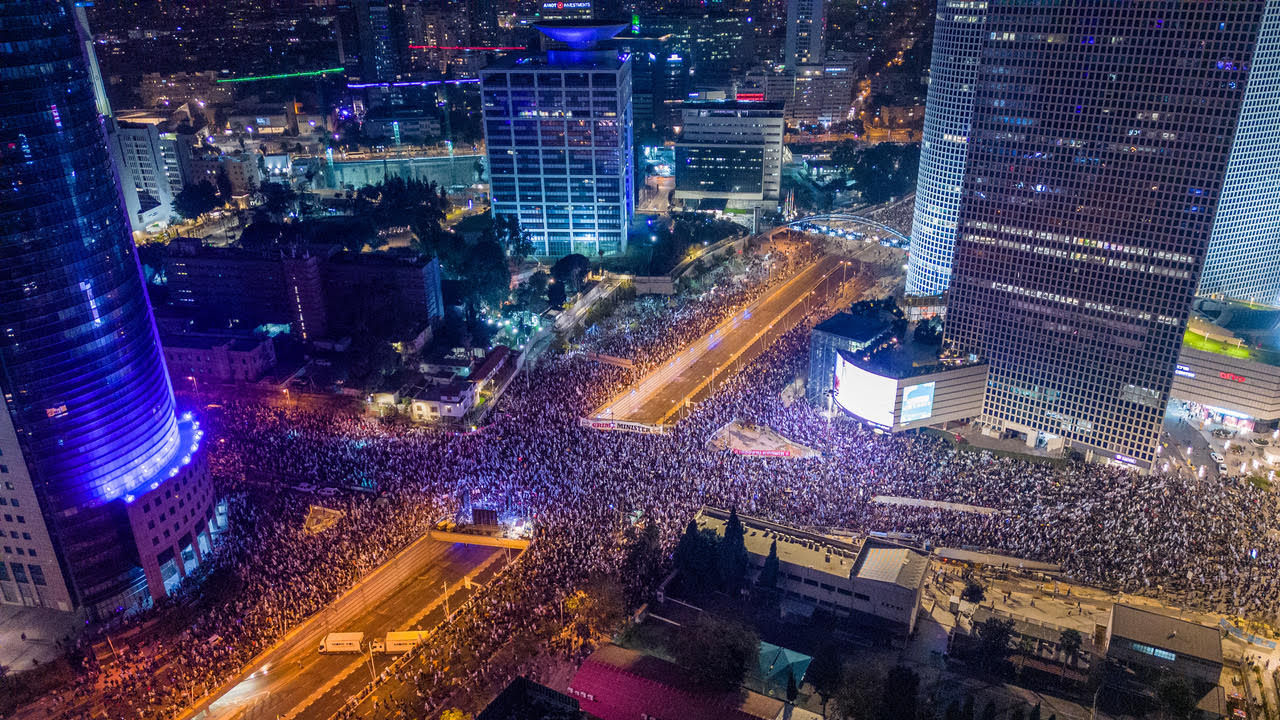 An aerial view of massive protests against the Israeli government's judicial overhaul legislation in Tel Aviv, Israel. You see massive crowds filling a major intersection between buildings.